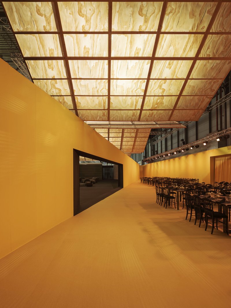 Corridor with ochre walls and backlit plywood ceiling grid leading to a dining area