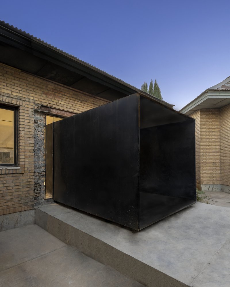 Black metal cube volume inserted between existing brick walls on a concrete plinth at twilight