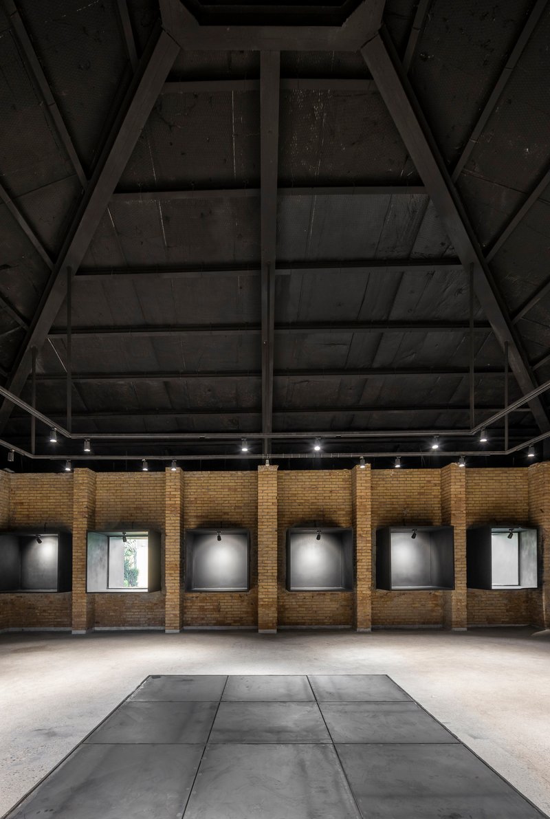 Interior gallery space with exposed timber roof trusses and framed artworks along yellow brick walls