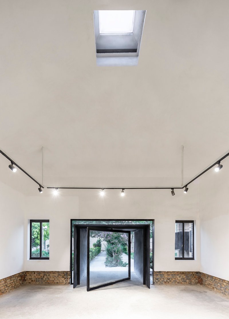 White-walled gallery with central pivot door, flanking windows, track lighting and a roof skylight above