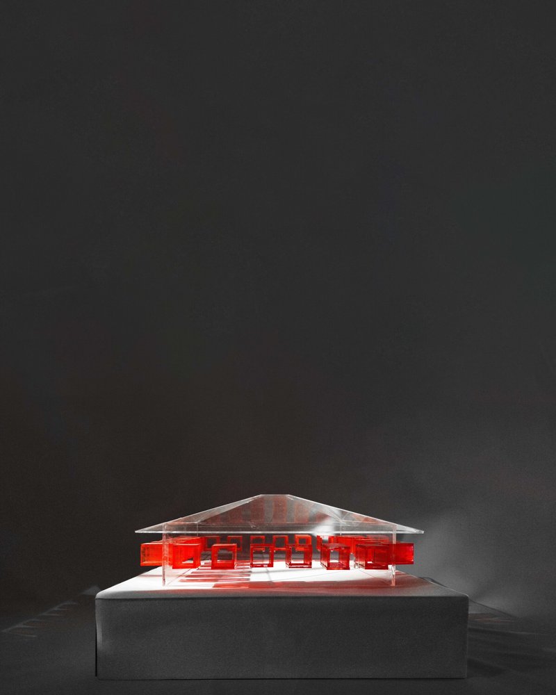 The image showcases an architectural model with a minimalist and modern design. The model features a transparent structure with red geometric accents, creating