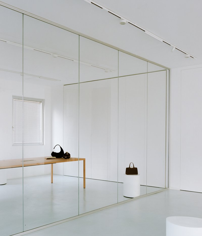 Floor-to-ceiling glass partition reflecting a timber bench and handbags with track lighting on the ceiling