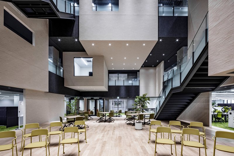 Multi-storey atrium with light wood-clad volumes, black ceilings and yellow chairs arranged on the ground floor