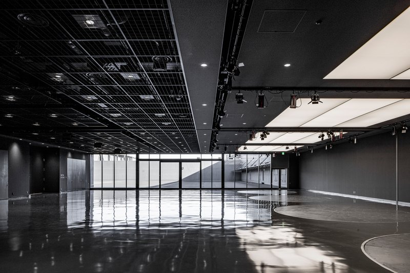 Open event space with black exposed ceiling grid and glazed walls overlooking an interior staircase