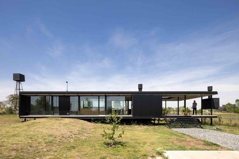 Elevated black corrugated metal pavilion with full-height glazing and covered terrace in open grassland