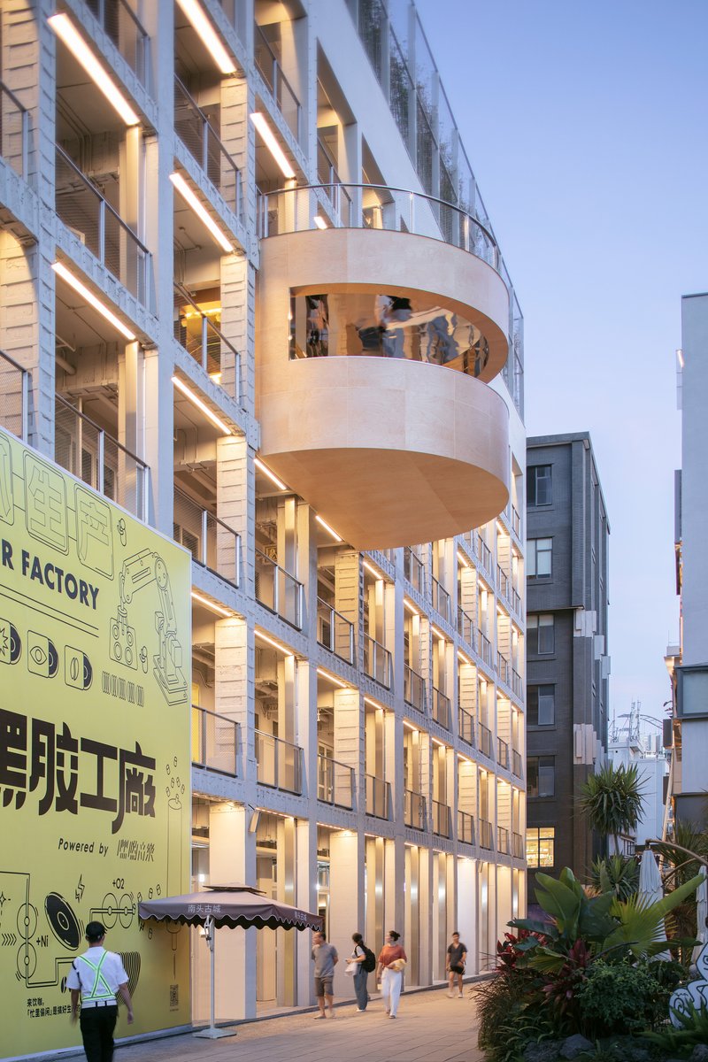 Curved balcony projecting from the illuminated facade of a multistory building at dusk with pedestrians below