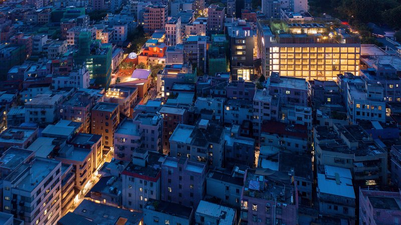 Aerial view of the illuminated glass facade rising above the surrounding dense low-rise neighborhood at dusk