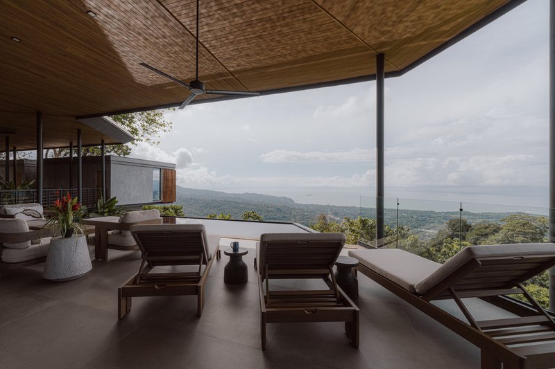 Covered terrace with timber ceiling and lounge chairs overlooking a forested valley in afternoon light