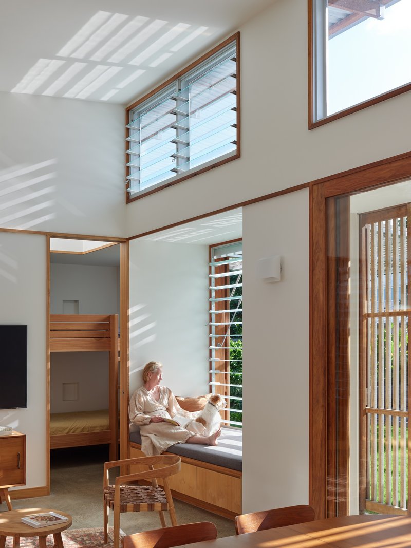 Double-height living area with woman seated on window bench beneath louvered windows in afternoon light