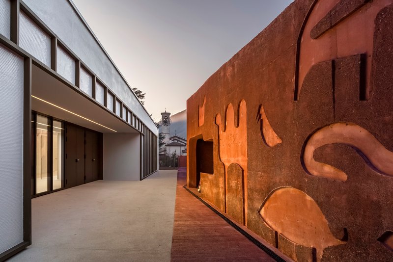 Courtyard passage between weathered steel sculptural wall and white volume with vertical glazing at dusk