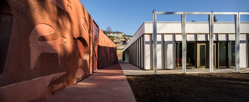 Exterior passageway with terracotta paving running between corten steel wall and white framed colonnade