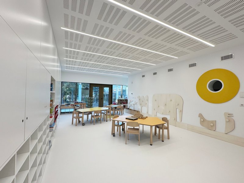 Classroom with plywood tables and chairs under perforated ceiling with linear luminaires and yellow circular wall element