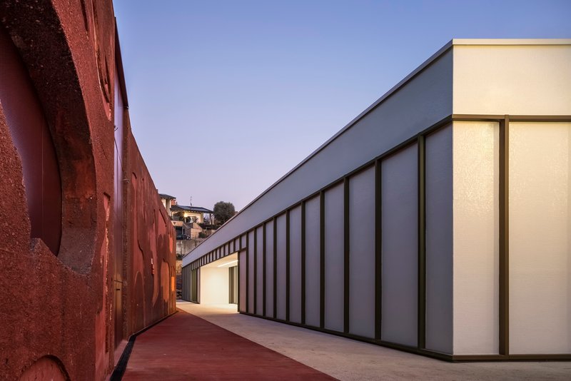 Narrow courtyard between the rammed earth wall and the translucent panel facade at dusk