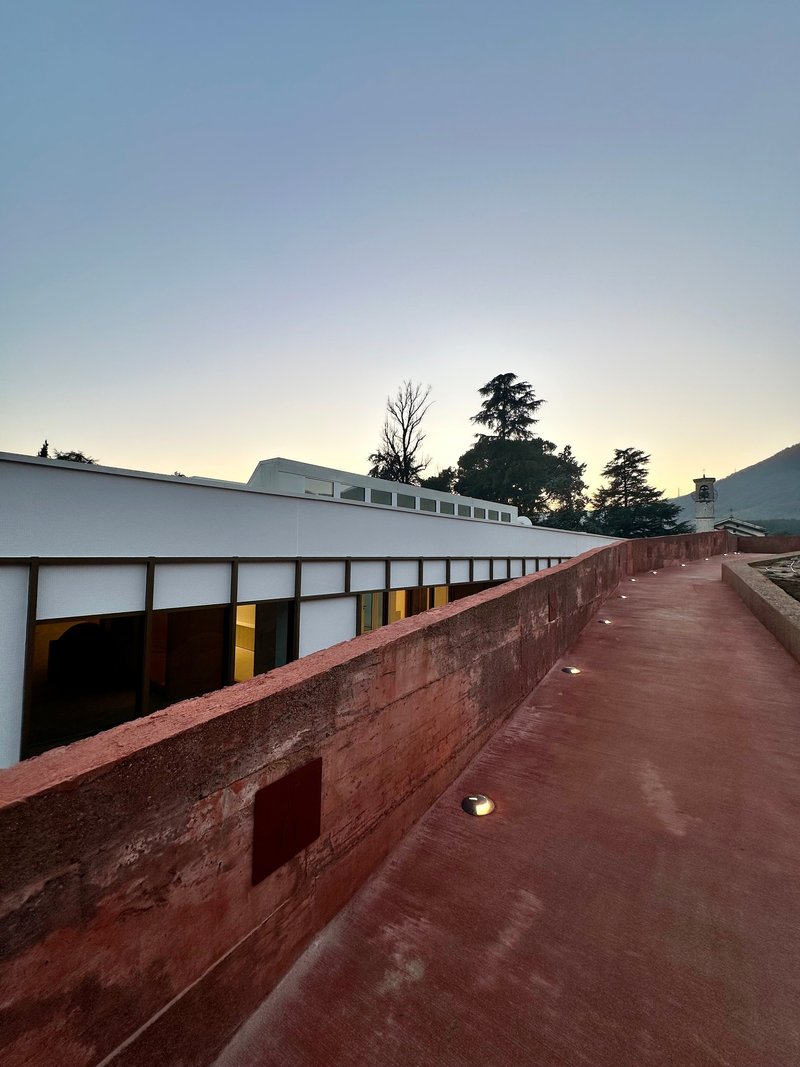 Rammed earth ramp ascending alongside the glazed facade under a pale twilight sky