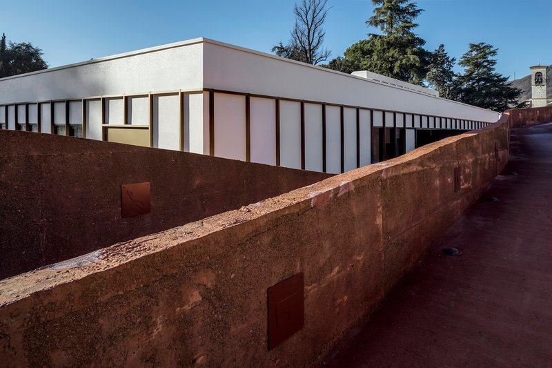 Long view of the white panel facade above the weathered rammed earth wall in daylight