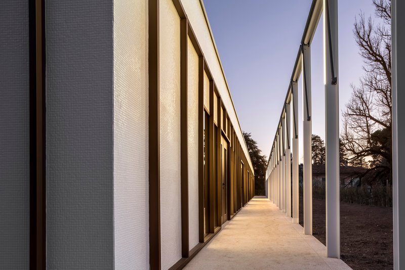 Covered walkway between textured panel walls with bare trees visible through the open side at sunset