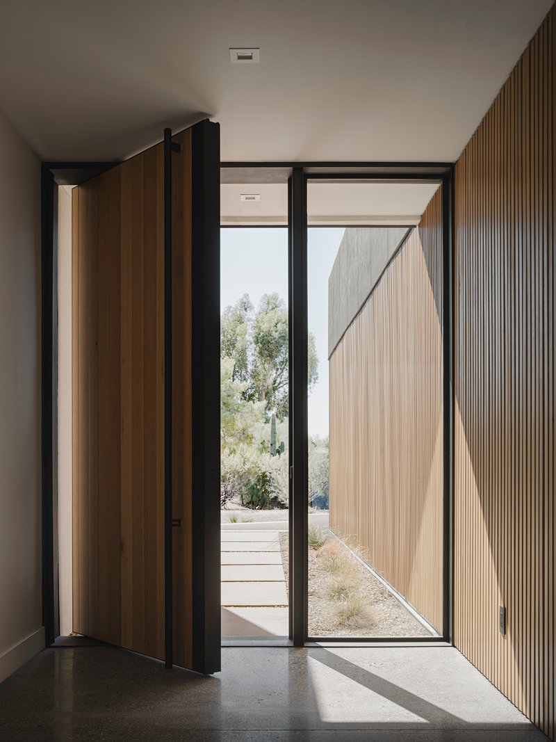Entry vestibule with pivoting timber door and vertical fluted wood wall panel casting shadows on polished concrete floor