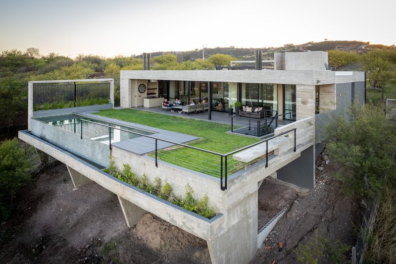 Elevated concrete residence with rooftop pool and lawn terrace surrounded by scrubland at dusk