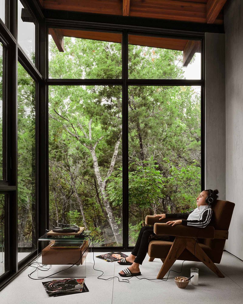 Interior seating nook with floor-to-ceiling windows framing trees where a person reclines listening to music