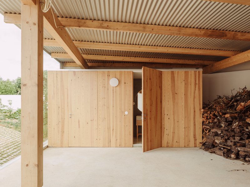 Covered carport with timber posts, exposed beams, corrugated metal ceiling, and sliding wood panel doors beside stacked firewood