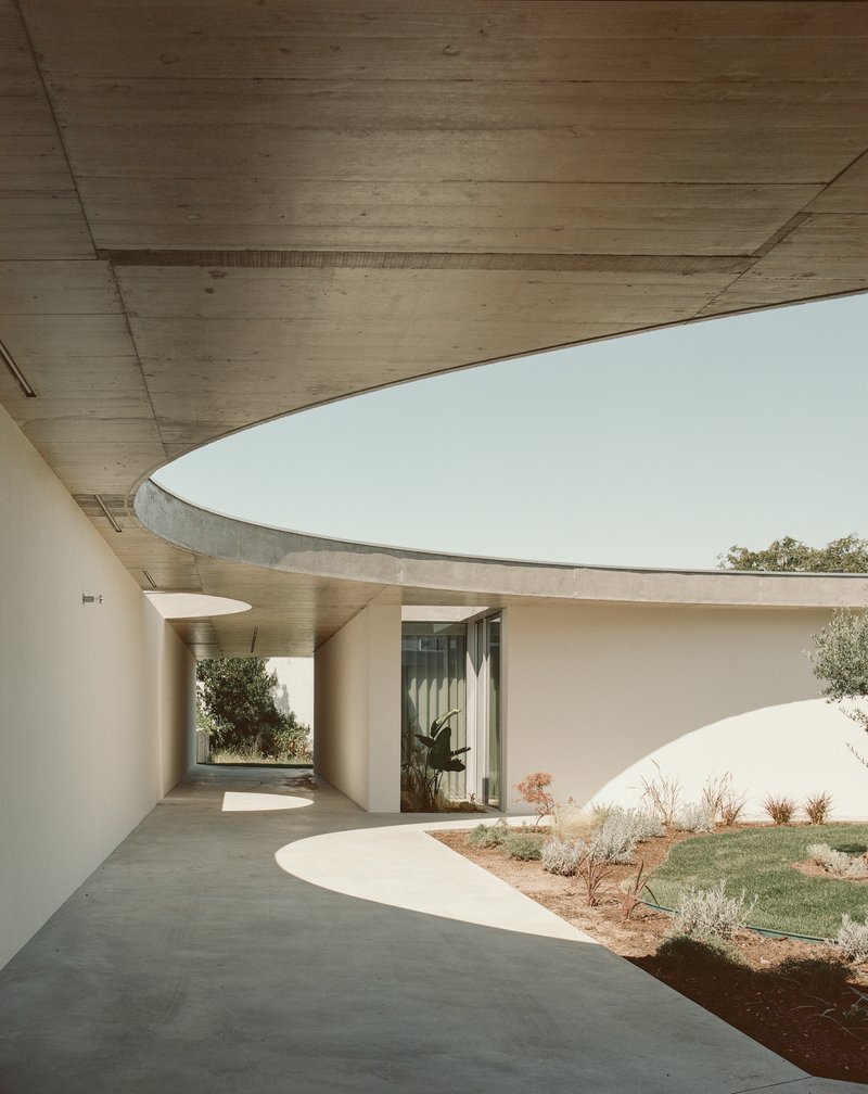 Curved covered walkway beneath exposed concrete ceiling with courtyard and plantings beyond
