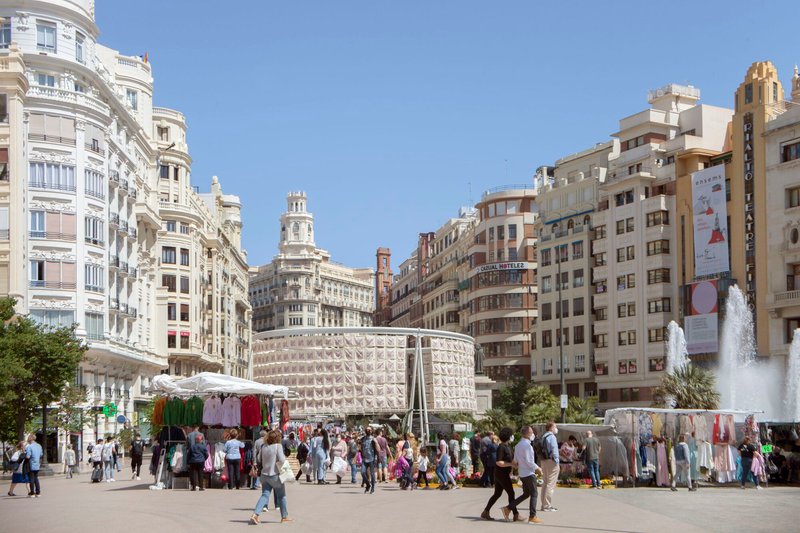 Temporary cylindrical pavilion with perforated white panels installed in a plaza surrounded by crowds and market stalls