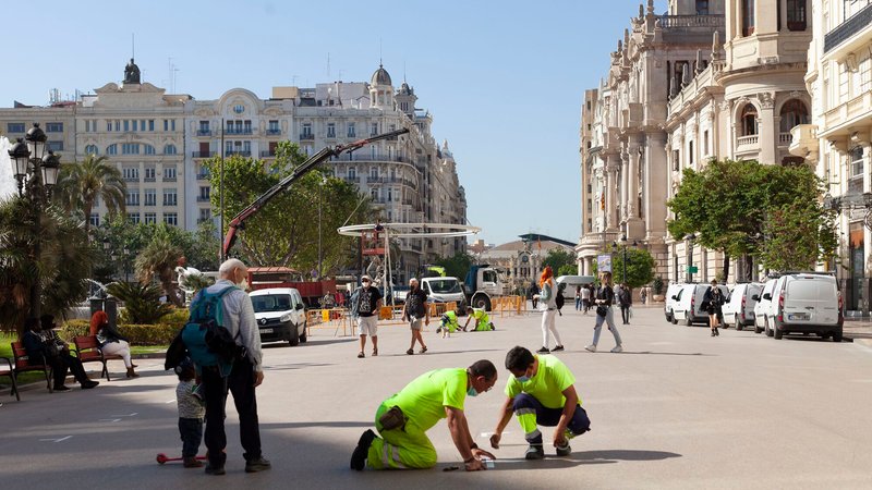 Construction crew measuring and marking positions on the plaza pavement as crane lifts structural element behind them