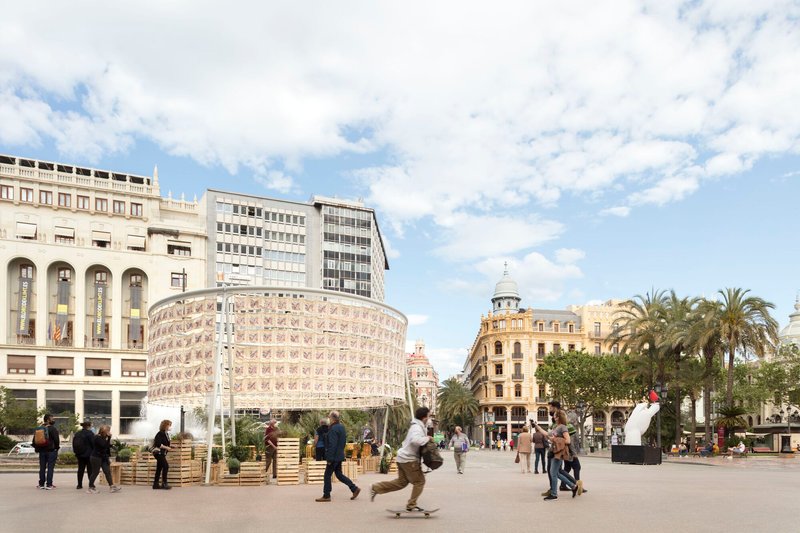 Cylindrical fabric pavilion with printed textile panels in a public plaza surrounded by palm trees