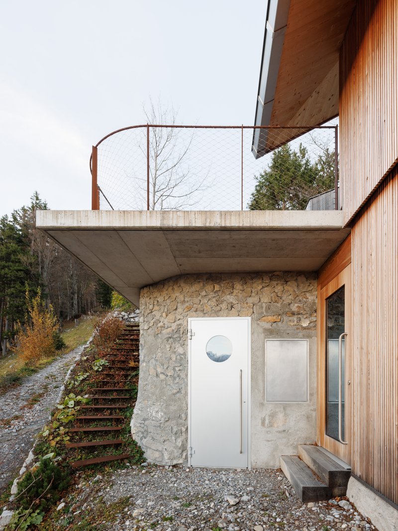 Entry door set into a stone base wall beneath a cantilevered concrete terrace with mesh railing