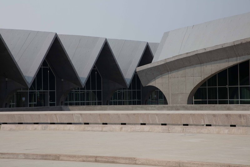 Concrete plaza with stepped seating facing a facade of zigzag gabled roofs and arched glass openings