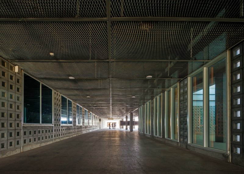 Long corridor with perforated metal ceiling and patterned concrete block walls flanking glazed openings