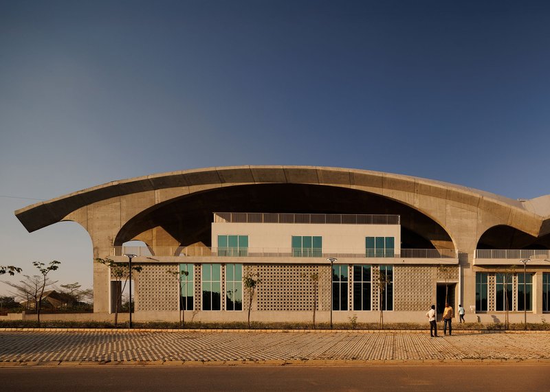 Curved concrete arches sheltering perforated brick facade with glazed openings at dusk with two figures on plaza