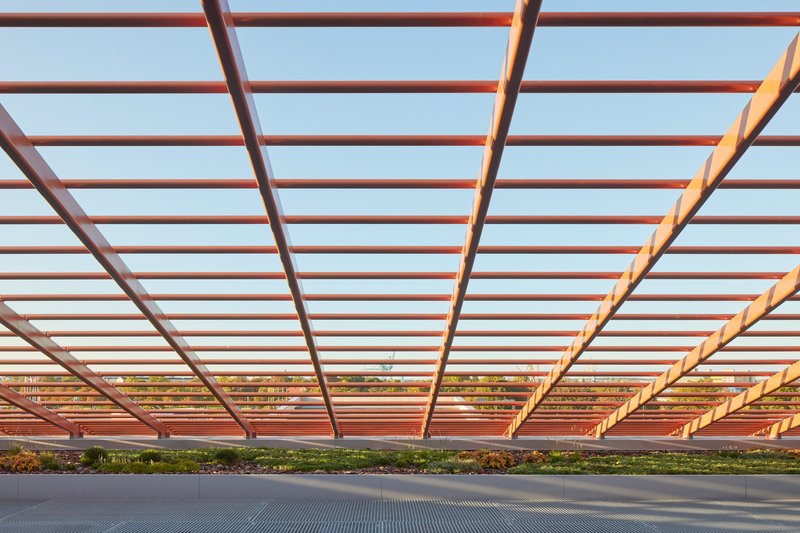 View beneath a pergola with red steel beams and glass infill panels at sunset