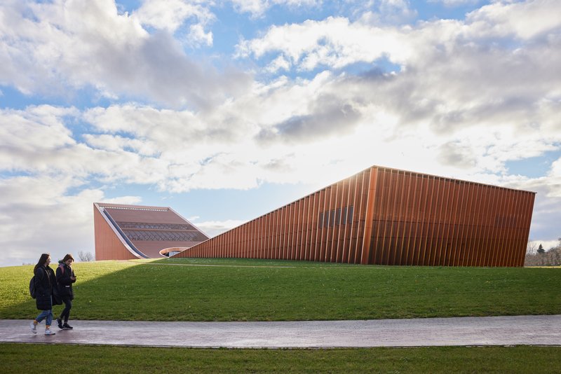 Vertical terracotta-clad facade emerging from a grassed berm with two figures walking along a path