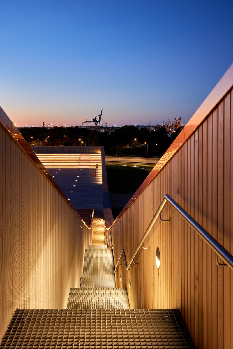 Exterior stairway descending between timber-clad walls with strip lighting at dusk