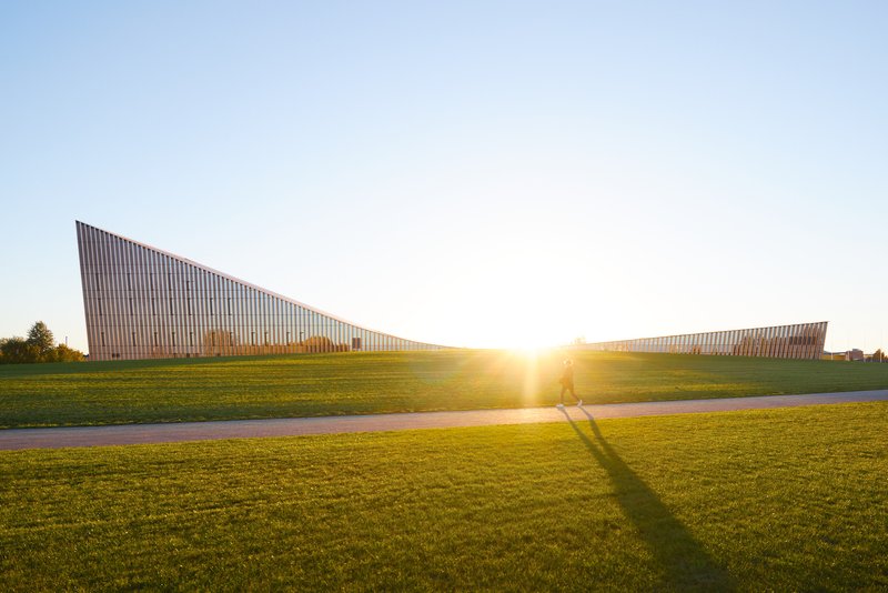 Two angled facade volumes rising from the grass lawn at sunrise with cyclists passing