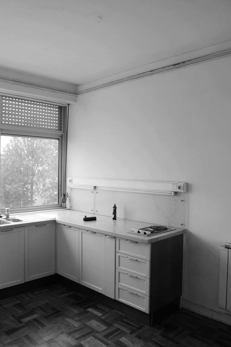 Corner kitchen with white cabinets and herringbone parquet flooring beneath a window with roller shutter