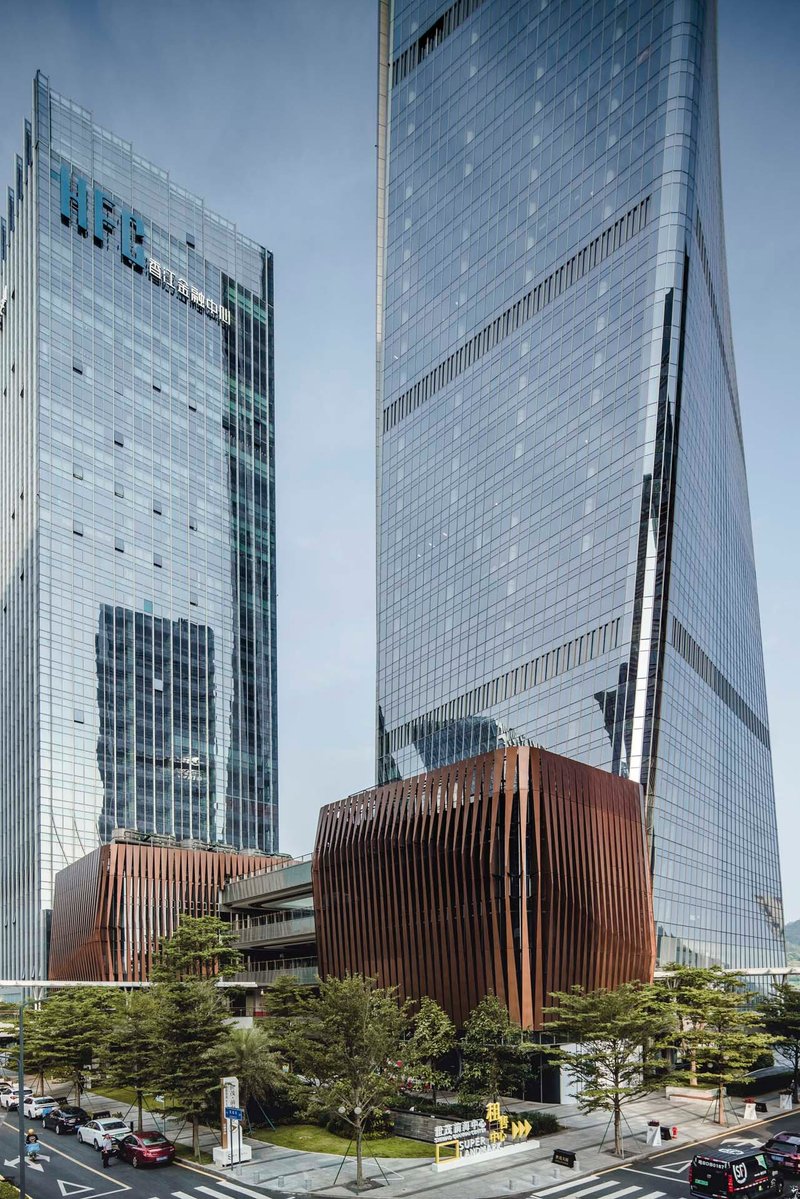 Curved bronze louvered pavilion at the base of the glass tower surrounded by planted evergreens