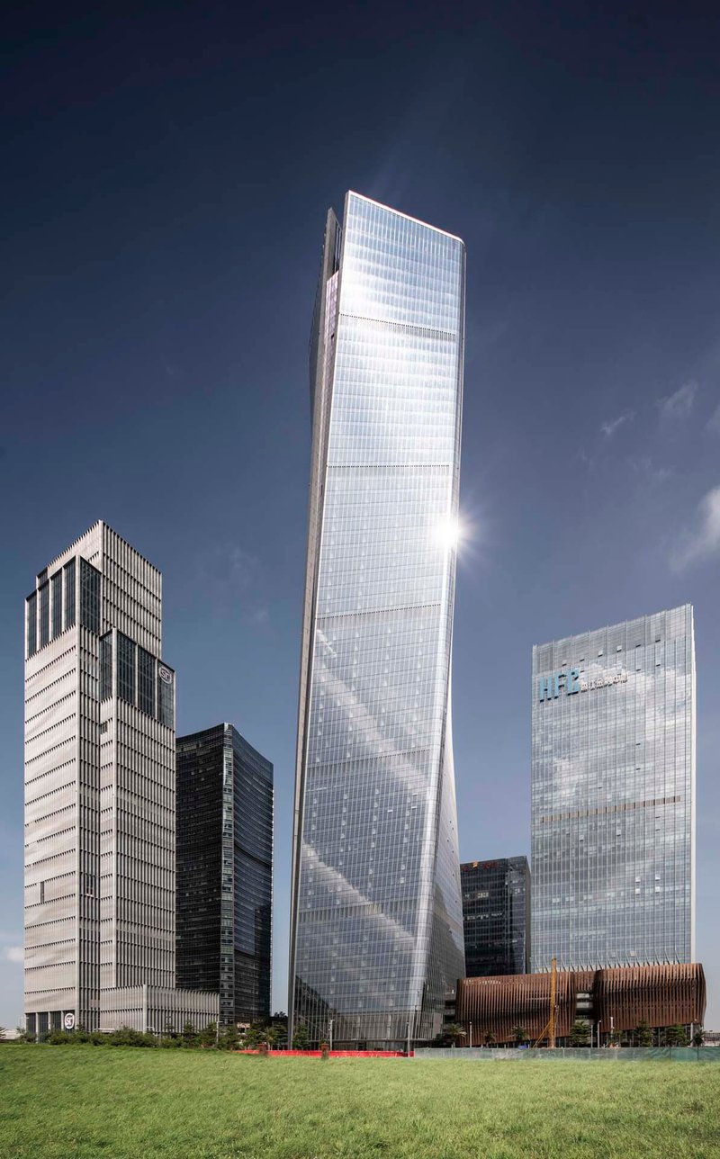 Twisted glass tower rising above surrounding buildings and a foreground lawn on a clear day
