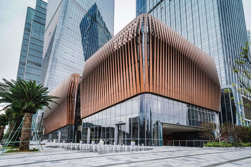Curved podium clad in vertical copper-toned fins above a glass base with fountain plaza and palms