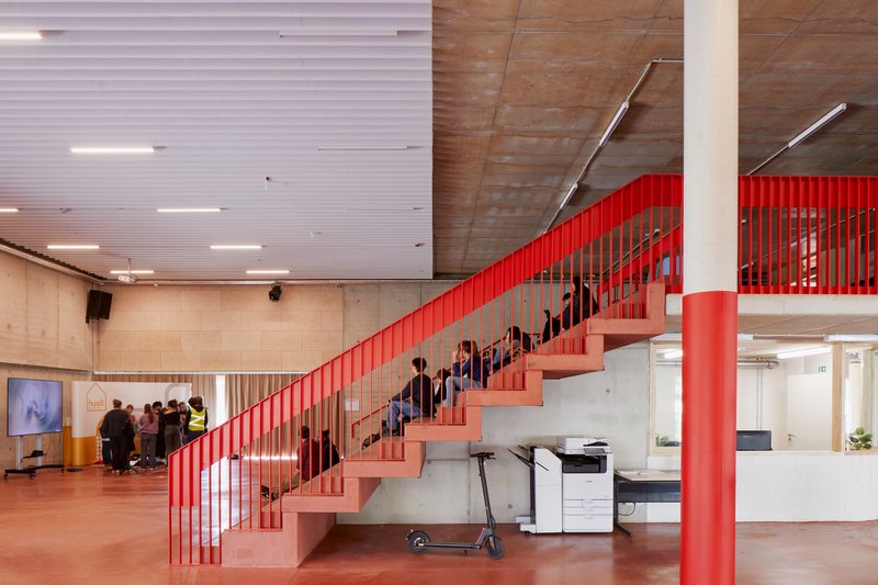 Interior atrium with red steel staircase, vertical balustrade, and concrete ceiling with fluorescent lighting above a gathering