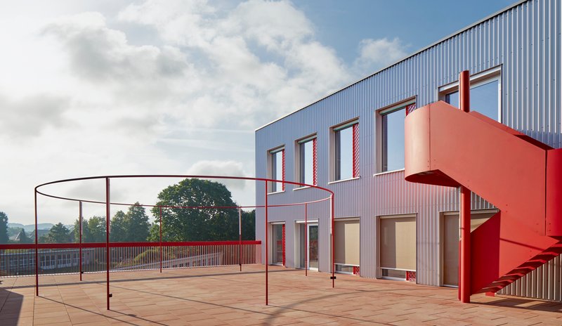 Rooftop terrace with red steel railing, play equipment, and external staircase beside corrugated metal facade