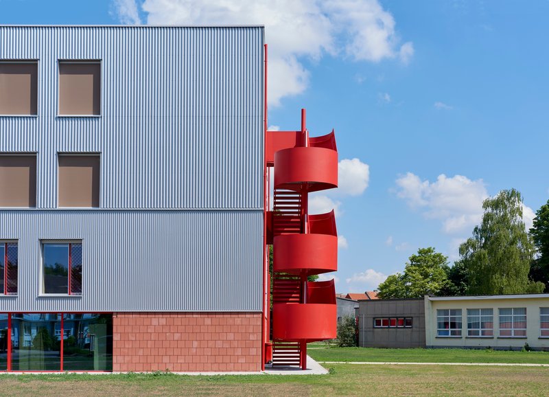 Close view of the red cylindrical staircase wrapping the corrugated metal facade under a blue summer sky