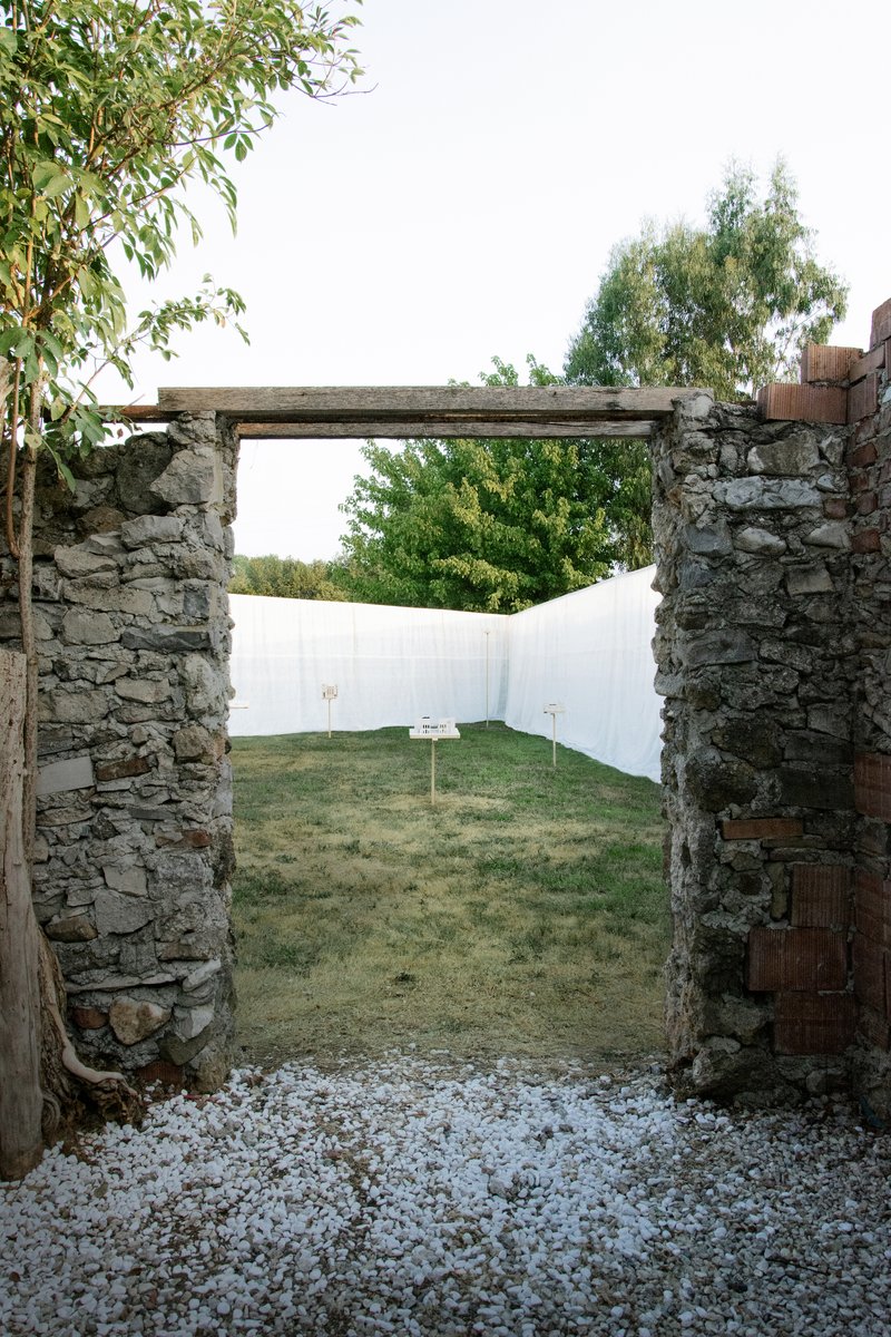 View through stone doorway with timber lintel toward white fabric wall and gravel path