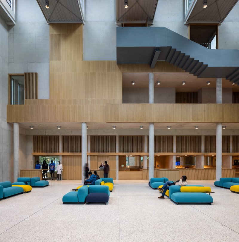 Lobby floor with timber service counter, concrete columns, and visitors seated on modular teal and yellow upholstered modules