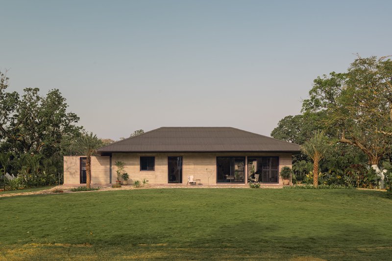 Rammed earth facade with hipped metal roof set against a manicured lawn and tree canopy at dusk