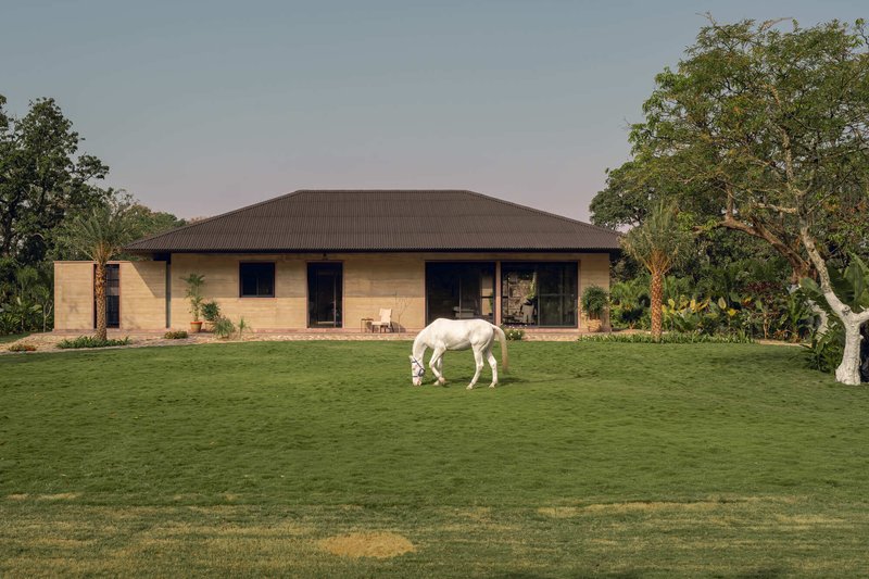 Rammed earth facade with hipped corrugated metal roof set in a lawn with a white horse grazing