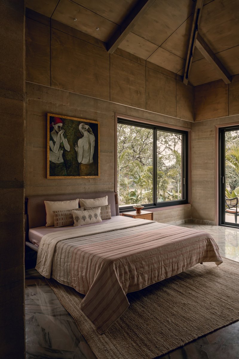 Bedroom with rammed earth walls and vaulted plywood ceiling overlooking trees through black-framed glazing