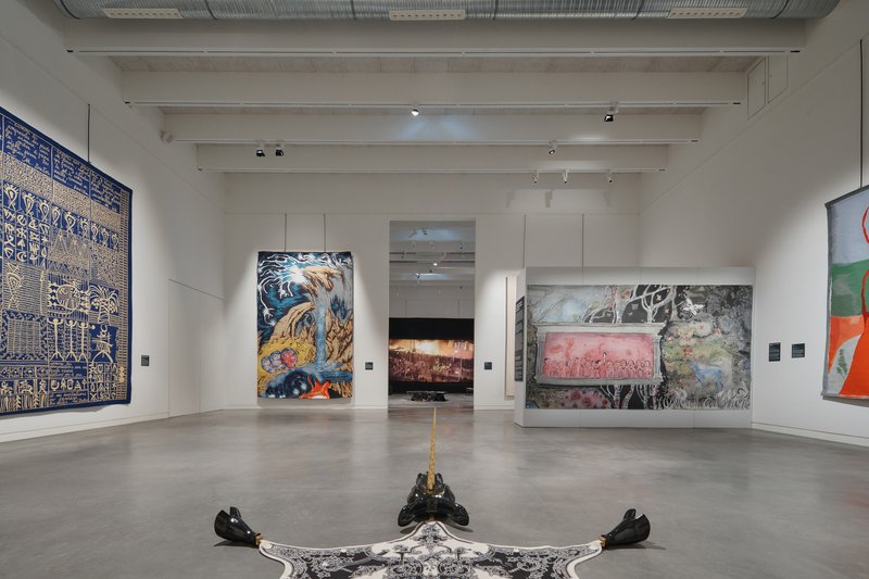 Exhibition hall with white ceiling beams and floor sculpture beneath hanging artworks arranged around doorways