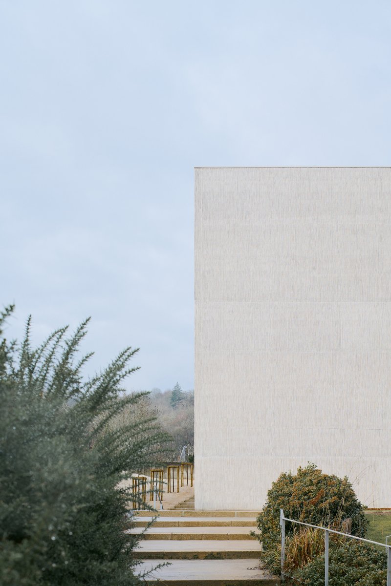 Concrete volume rising above wide exterior steps framed by planted shrubs under a pale sky